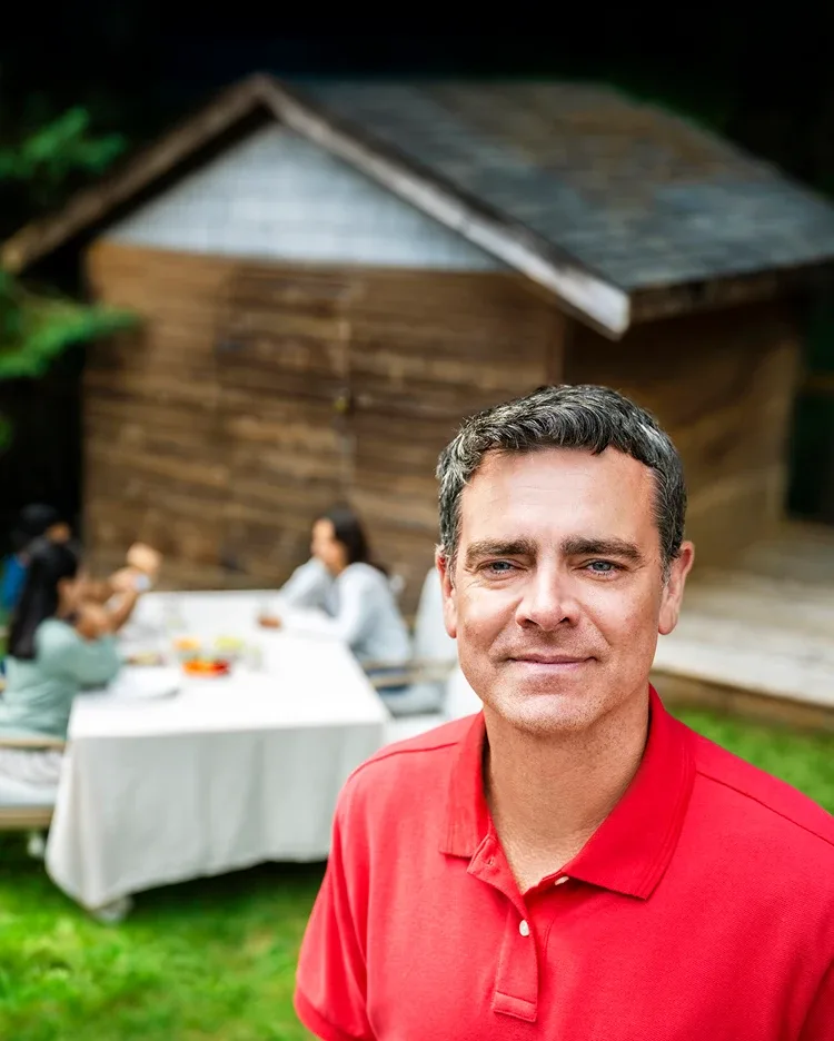 A man smiling and standing outdoors and people are sitting at a table in the background